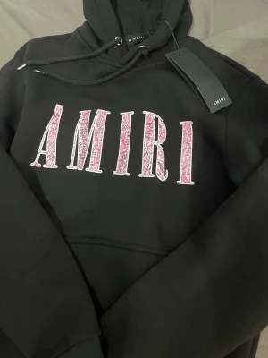 Svart Amiri hoodie - Snygg svart hoodie från Amiri med stort rosa och vitt AMIRI-tryck på bröstet. Klassisk huva med dragsko och känguruficka framtill. Perfekt för dig som gillar streetwear och vill sticka ut med ett exklusivt plagg.