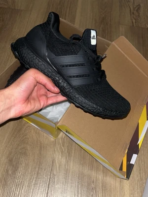 Svarta Adidas Ultraboost sneakers - Snygga helsvarta Adidas Ultraboost sneakers med stickad ovandel och klassiska tre ränder på sidan. Skon har en chunky sula med Boost-dämpning för extra komfort och en sportig look. Perfekt för dig som gillar streetwear och vill ha något stilrent.