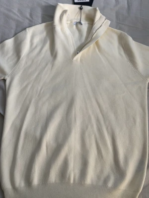 Off white halvzip tröja Tour & Tate - Snygg off white stickad tröja från Tour & Tate med halvzip och hög krage. Tröjan har ribbade muddar och är tillverkad i mjukt material som känns skönt mot huden. Perfekt för lager på lager och enkel att styla till olika outfits.