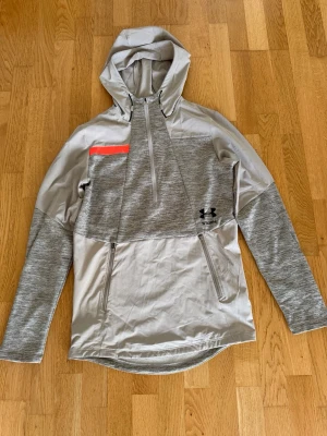 Grå hoodie från Under Armour - Snygg beige hoodie från Under Armour med halv dragkedja, huva och två dragkedjefickor. Tröjan har en diskret orange detalj på bröstet och är tillverkad i ett funktionsmaterial som passar perfekt för träning eller bara chilla