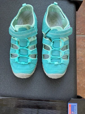 Turkosa sportiga sandaler med kardborre - Säljer ett par turkosa sandaler med sportig design och kardborreband. De har stängd tå, öppna sidor och elastiska snören framtill. Materialet är en mix av textil och syntet, med vadderad innersula för extra komfort. Perfekta för sommardagar och äventyr. Kolla gärna mina andra annonser❗️