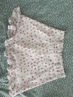 Vit blommig kortkjol från SHEIN - Söt vit kortkjol från SHEIN med små rosa blommor och volangkant. Kjolen har en diskret dragkedja bak och är gjord i ett lätt, luftigt material som passar perfekt till varma dagar.