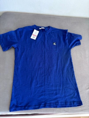 Blå t-shirt från Calvin Klein - Snygg blå t-shirt från Calvin Klein med klassisk passform och rund hals. T-shirten har korta ärmar och en liten CK-logga broderad på bröstet. Perfekt för en stilren och enkel look.