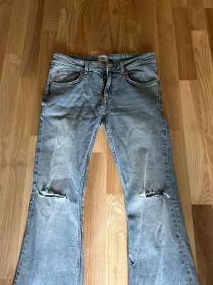 Lågmidjade Ljusblå slitna bootcut jeans - Ljusblå jeans med bootcut passform och slitningar på knäna. Perfekt för en avslappnad och trendig look. Använda fåtal gånger och vill bli av med. Köpta för 400 