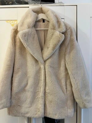 Beige fluffig pälsjacka - Säljer en supermjuk och fluffig beige pälsjacka i fuskpäls. Jackan har bred krage, raka ärmar och en klassisk rak passform. Perfekt för dig som vill ha en varm och trendig look under kalla dagar. Stängs framtill och har en stilren design.