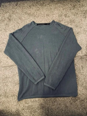 Blå tunn stickad tröja - En enkel och stilren blå stickad tröja med lång ärm och rund hals. Tröjan har en ribbad struktur och är perfekt för lager på lager. Passar dig som gillar en clean och avslappnad look. Detaljerad stickad med ett snyggt mönster. 