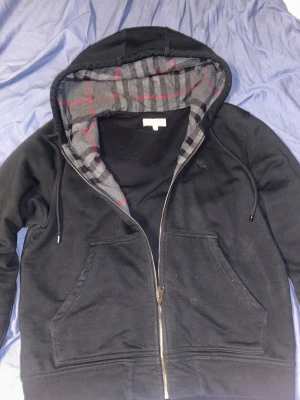 Svart zip hoodie från burberry - Svart burberry zip hoodie med dragkedja och huva. Insidan av huvan och öppningen är fodrad med grått tyg i rutigt mönster med röda och svarta linjer. Två stora fickor framtill och justerbar dragsko i huvan. Perfekt för en chill och avslappnad stil.