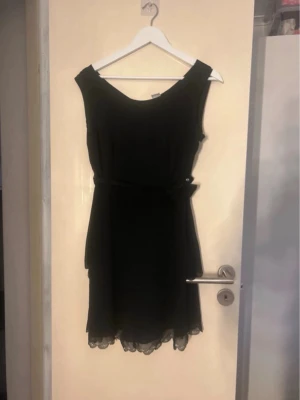 Svart kort klänning med spetsdetaljer - Stilren svart kort klänning med spetsdetaljer längst ner och knyt i midjan. Klänningen är ärmlös och har en mjuk, följsam passform som ger en elegant siluett. Perfekt för dig som gillar klassisk och enkel stil.