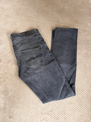 Nudie jeans grim tim - Säljer ett par mörkgrå jeans med rak passform och klassiska fem fickor. Jeansen har snygga sömdetaljer på bakfickorna och är tillverkade i ett mjukt denimtyg. Perfekta för en avslappnad och stilren look. Grim Tim - 32/32