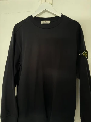 Svart sweatshirt från Stone Island - Svart långärmad sweatshirt från Stone Island med klassisk rund halsringning och ikonisk logotyp-patch på vänster ärm. Tröjan har en clean look och är tillverkad i mjukt bomullsmaterial som känns skönt mot huden.