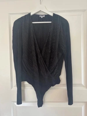 Svart plisserad body med omlott - Snygg svart body med plisserat tyg och omlott-ringning. Långa ärmar och åtsittande passform nedtill. Materialet ser ut att vara mjukt och stretchigt, perfekt för att skapa en cool och trendig look. Passar till både jeans och kjol.