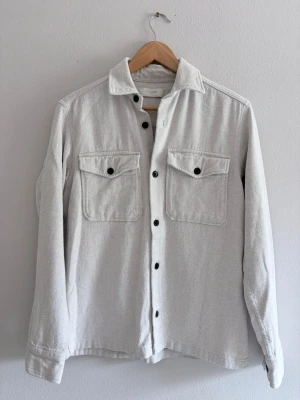 Ljusgrå overshirt från Jack & Jones - Säljer en stilren ljusgrå overshirt från Jack & Jones Premium. Skjortan har två bröstfickor med knappar, klassisk krage och svarta knappar framtill. Perfekt lager-på-lager-plagg med lång ärm och avslappnad passform.