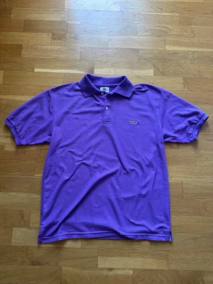 Lila Lacoste Piké - En mycket fin och stilren lila piké från Lacoste.  Storlek: 5/L Passform: Normal Skick: Fint skick utan några hål eller fläckar, något nopprig (se bilder noga)!  Har du några frågor eller vill ha fler bilder? Tveka inte på att höra av dig!