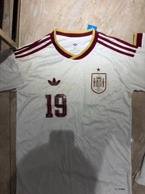 Vit Spanien fotbollströja nr 19 - Säljer en vit Spanien fotbollströja från Adidas med vinröda detaljer, nummer 19 och namnet Lamine Yamal på ryggen. Tröjan har gula och vinröda ränder vid kragen och axlarna samt broderat landslagsemblem på bröstet. Perfekt för fotbollsfans!