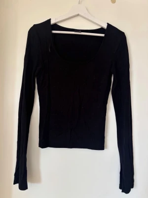 Svart långärmad topp med vid urringning - En enkel och stilren svart långärmad topp med vid, rund urringning. Modellen är figurnära och har långa ärmar. Materialet ser ut att vara mjukt och stretchigt, perfekt för lager på lager eller som basplagg.