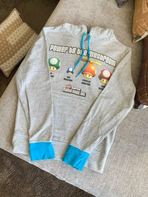 Grå Super Mario Bros. Wii hoodie - Säljer en grå hoodie med färgglada Super Mario-svampar och texten 'Power to the Mushroom' på bröstet. Hoodien har blåa muddar och dragsko, samt en stor tryckt logga från New Super Mario Bros. Wii. Perfekt för dig som älskar Nintendo och gaming.
