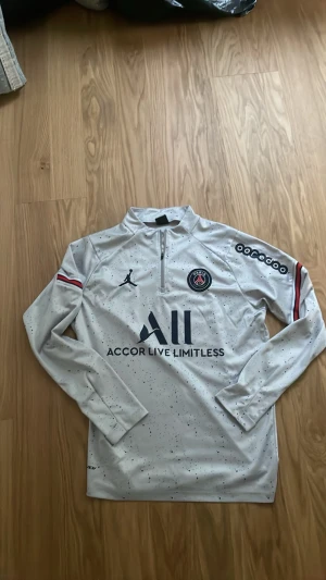Vit PSG träningströja med halv dragkedja - Säljer en vit långärmad PSG träningströja med svarta och röda detaljer. Tröjan har halv dragkedja framtill, PSG-logga på bröstet och texten 'Accor Live Limitless'. Materialet är troligtvis polyester, perfekt för träning eller fotboll.