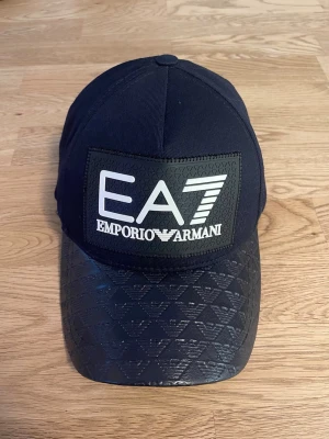 Svart keps EA7 - Snygg svart keps från EA7 med stort logotypmärke framtill och mönstrad skärm med diskreta loggor. Justerbar baktill med metallspänne. 