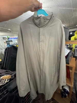 Beige minimalistisk trenchcoat - Stilren beige trenchcoat med hög krage och dold dragkedja framtill. Jackan har raka ärmar och två sidofickor. Passar perfekt för dig som gillar en enkel och clean look. Materialet ser ut att vara ett tunt tyg, troligtvis polyester eller bomullsblandning. Finns att hämta i Gröndal.