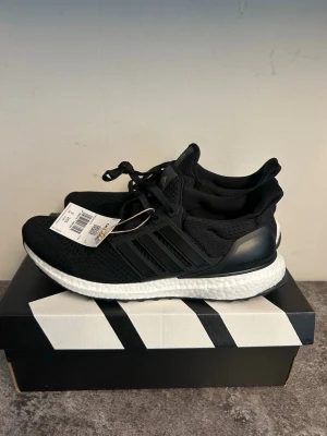 Adidas Ultraboost svarta sneakers - Svarta Adidas Ultraboost sneakers med stickad ovandel och vita Boost-sulor. Skorna har klassiska tre ränder på sidan, snörning och bekväm passform. Perfekta för dig som gillar sportig och stilren look.
