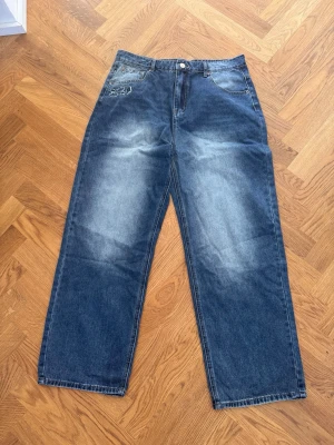 Derschutze jeans - Säljer ett par blå baggy jeans med ljusa slitningar och broderade detaljer på fickorna. Jeansen har raka ben och klassisk femficksmodell. Perfekta för en avslappnad och trendig stil.