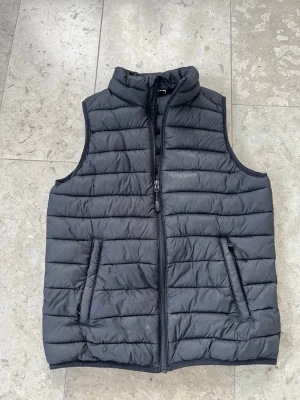 Svart dunväst - Snygg svart dunväst. Västen har dragkedja framtill och två fickor med dragkedja. Perfekt att slänga över en hoodie eller tröja för extra värme. Materialet är lätt och smidigt, vilket gör den enkel att bära.
