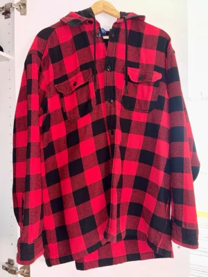 Röd- och svartrutig skjorta med huva - Overshirt i röd- och svartrutigt mönster med svarta knappar framtill och två bröstfickor med lock. — Modellen har en svart huva med dragsko och är gjord i ett mjukt flanellmaterial. — Perfekt för lager på lager och en avslappnad streetwear-look. — En av knapphålen är aningen trasig, men plagget är annars i bra skick. — Nypris ca. 300 kr.