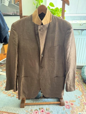 Loro Piana kavaj - Snygg brun kavaj från Loro Piana med unik beige krage i suede. Kavajen har 4 knappar framtill, tre fickor och en modern, rak passform. Materialet är en blandning av ull och bomull, vilket ger en exklusiv känsla.