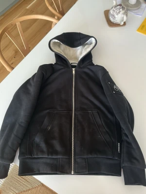 Äkta Svart hoodie från Moose Knucklers - Säljer en svart hoodie från Moose Knucklers med dragkedja framtill och två stora fickor. Hoodien har en mjuk, ljus insida i huvan och ribbade muddar vid ärmar och nederkant. Detalj med logga på ena ärmen och klassisk streetwear-look.