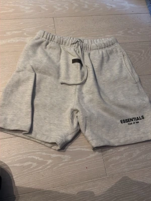Grå shorts Essentials Fear of God - Säljer ett par ljusgrå shorts från Essentials Fear of God med tryck på benet. Shortsen har resårmidja med snörning och är tillverkade i mjuk bomullsmix. Perfekta för chill dagar eller streetwear-stil.