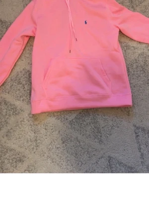 Rosa hoodie med huva och ficka - En snygg rosa hoodie med huva och dragsnören samt en stor magficka. Liten blå logga broderad på bröstet. Hoodien har långa ärmar och ribbade muddar vid ärmslut och nederkant. Perfekt för en avslappnad och trendig look.