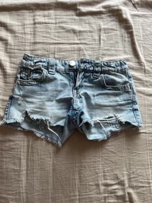Ljusa jeansshorts med slitna detaljer - Säljer ett par ljusblå jeansshorts med fransiga kanter och slitna detaljer framtill. Shortsen har snygg broderad design på bakfickorna. Perfekta för sommardagar och enkel att matcha med allt.