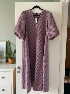 Rutig långklänning med puffärm Lindex - Långklänning i rosa och lila rutigt mönster med puffiga kortärmar och v-ringad hals. Klänningen har en avslappnad passform och  materialet är i bäckebölja. Perfekt för dig som gillar en trendig och bekväm stil.