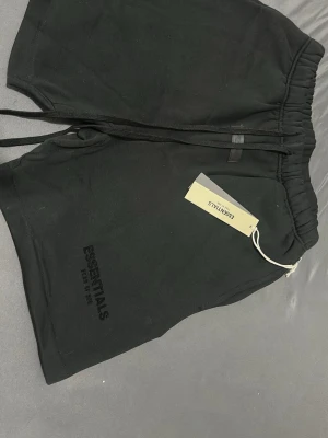 Svarta shorts från Essentials - Svarta shorts från Essentials med elastisk midja och snörning. Klassisk och enkel design med diskret logga på benet. Perfekta för en avslappnad stil och tillverkade i mjukt material som känns skönt mot huden.