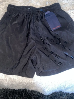 Svarta shorts från Louis Vuitton - Snygga svarta shorts från Louis Vuitton med klassisk logga i blankt tryck på ena benet. Shortsen har elastisk midja med snörning och är tillverkade i ett lätt, syntetiskt material som passar perfekt till sommaren.