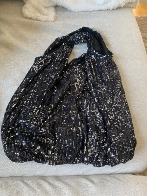 Svart paljettväska  - Säljer en svart handväska täckt av glittrande paljetter. Väskan har en mjuk, slouchy form och breda axelband. Materialet är tyg med paljettöverdrag som ger en riktigt festlig look. Perfekt för dig som vill sticka ut och addera lite extra bling till din outfit.