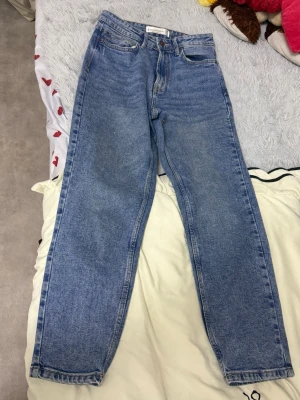 Blå mom jeans från Stradivarius - Säljer ett par klassiska blå mom jeans från Stradivarius i storlek 38. Jeansen har hög midja, raka ben och fem fickor. Tillverkade i ett slitstarkt denimtyg med snygg tvättad look och kontrastsömmar. Perfekta för en avslappnad och trendig stil.