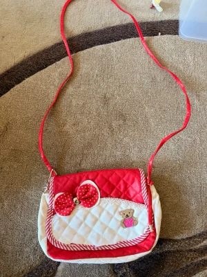 Röd och vit axelväska med rosett - Söt axelväska i quiltad röd och vit design med en stor prickig rosett och en liten björn-detalj framtill. Väskan har ett långt axelband💞