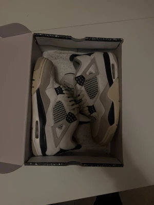 Air Jordan 4 Retro - Säljer ett par gråa Air Jordan 4 Retro sneakers med svarta och vita detaljer. Skorna har meshpaneler, synlig Air-sula och klassisk Jumpman-logga på plösen. Stilren design med coola kontraster och robust känsla. Perfekta för dig som gillar streetwear och sneakerskultur.