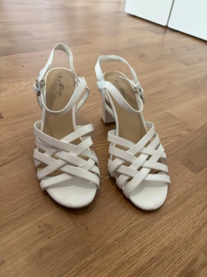 Vita klackade sandaler med remmar - Säljer ett par vita sandaler med bred klack och flätade remmar över foten. Sandalerna har öppen tå, justerbart spänne vid ankeln och en stabil blockklack. Perfekta för sommaren och ger en stilren look till din outfit.