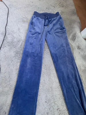 Blå velour mjukisbyxor med dragsko - Säljer ett par blå mjukisbyxor i mjuk velour med bred resår i midjan och dragsko. Byxorna har raka ben och två stora fickor framtill. Perfekta för chill dagar eller när du vill vara extra bekväm.