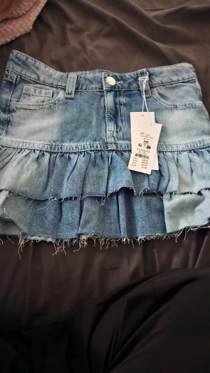 Blå jeanskjol med volanger - Säljer en blå jeanskjol med volangdetaljer och råa kanter. Kjolen har klassisk femficksdesign, knapp och dragkedja framtill samt bälteshällor. Perfekt för dig som gillar en avslappnad och trendig stil.