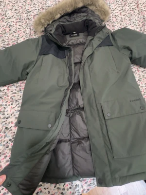 Jacka - Stilren grön parkas från Everest med svarta axelpartier och avtagbar huva med fuskpäls. Jackan har stora fickor med knappar, dragkedja och tryckknappar framtill samt reflekterande detalj bak. Perfekt för kalla dagar. Passar både S-M kan också passa L faktiskt