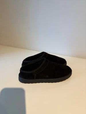 Svarta slip-in tofflor i mocka - Ett par svarta slip-in tofflor med mjuk ovandel i mocka och fodrad insida. Klassisk och enkel design med rund tå och låg sula. Perfekta för att chilla hemma och hålla fötterna varma.
