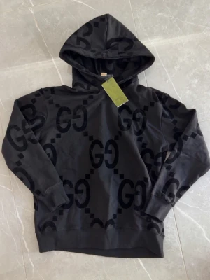 Svart hoodie med GG-mönster från Gucci - Snygg svart hoodie från Gucci med stort GG-mönster över hela tröjan. Hoodien har en klassisk huva och ribbade muddar vid ärmar och nederkant. Tillverkad i mjukt material som känns skönt mot huden. Perfekt för dig som gillar exklusiv streetwear.