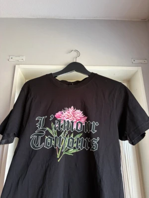 Svart t-shirt med blomtryck - Svart t-shirt med trycket 'L'amour Toujours' och en stor rosa blomma framtill. Klassisk rund hals och korta ärmar. Perfekt för dig som gillar statement-plagg med coolt tryck och lite romantisk vibe.