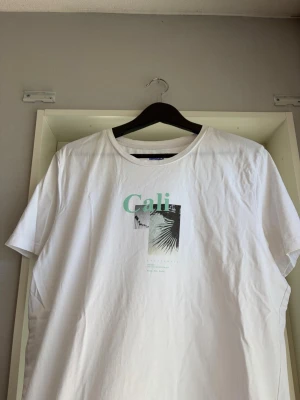 Vit t-shirt med Cali-tryck - Vit t-shirt med tryck framtill där det står 'Cali' i mintgrönt och motiv av palmer och blad. Klassisk rund halsringning och korta ärmar. Perfekt för en avslappnad stil.