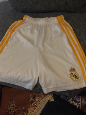 Vita Real Madrid shorts med gula ränder - Säljer ett par vita sportshorts med gula ränder längs sidorna och Real Madrid-logga på ena benet. Shortsen har resår i midjan och är tillverkade i ett lätt och snabbtorkande material, perfekta för träning eller match.