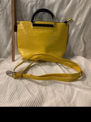 Gul croco handväska från Don Donna - Snygg dovt gul handväska från Don Donna i croco-mönstrat konstläder. Väskan har ett svart handtag i plast/metall och en lång axelrem som går att ta av. Perfekt för dig som vill sticka ut med färg och cool struktur.