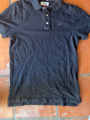 Marinblå pikétröja Tommy Jeans S - Klassisk mörkblåpikétröja från Tommy Jeans i slim fit-modell. Tröjan har krage, knappar framtill och en liten broderad logga på bröstet. 